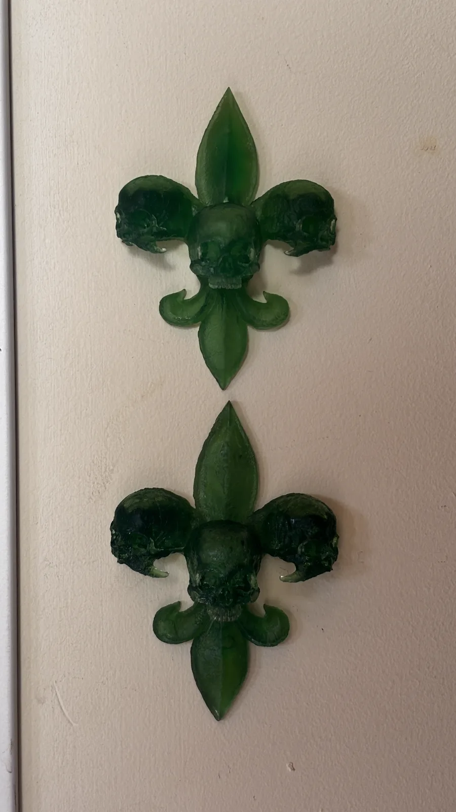 A fleur-de-lis built from skulls, cast in deep red translucent resin, mounted on a pale wall with its shadow beside it