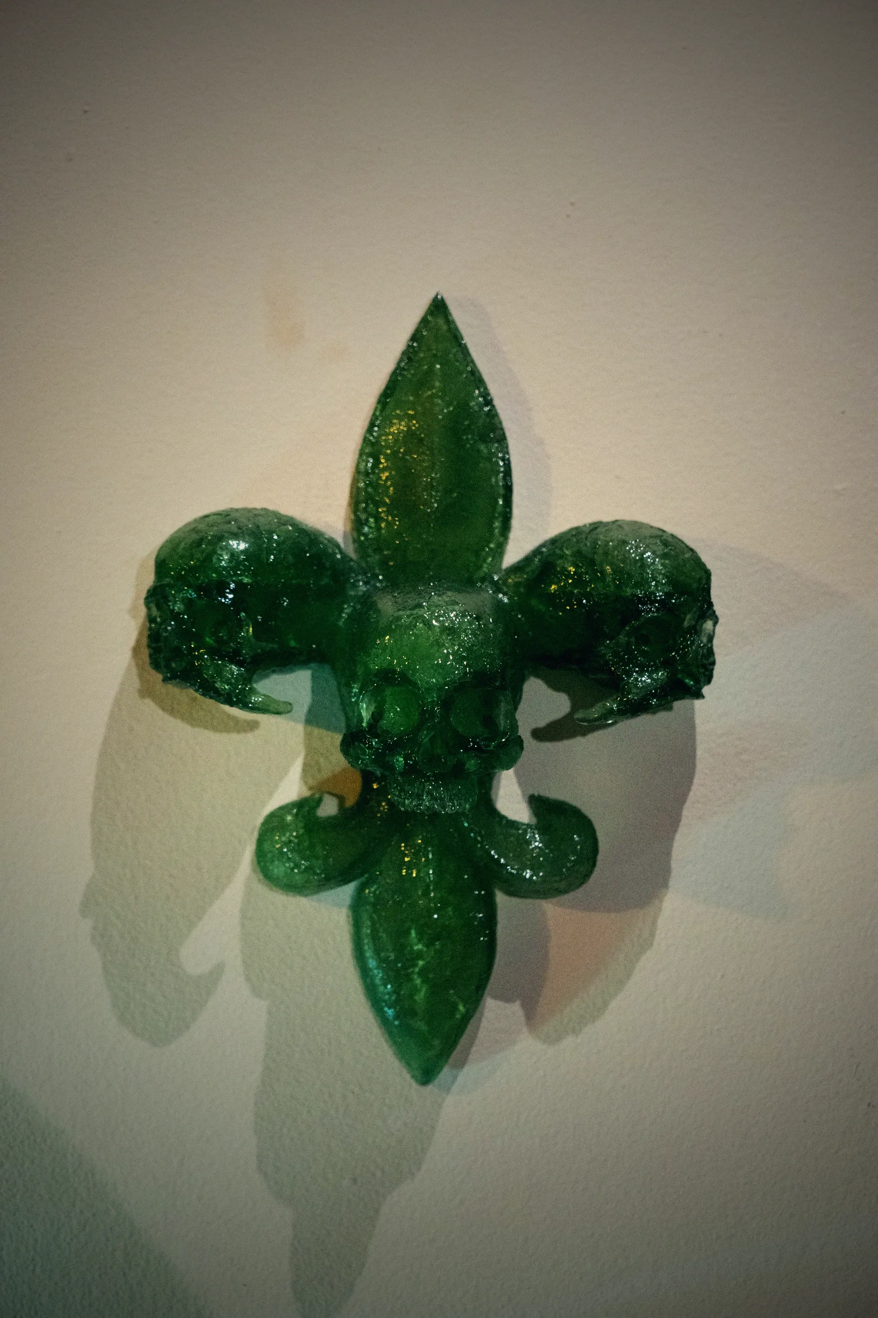 A fleur-de-lis built from skulls, cast in deep red translucent resin, mounted on a pale wall with its shadow beside it