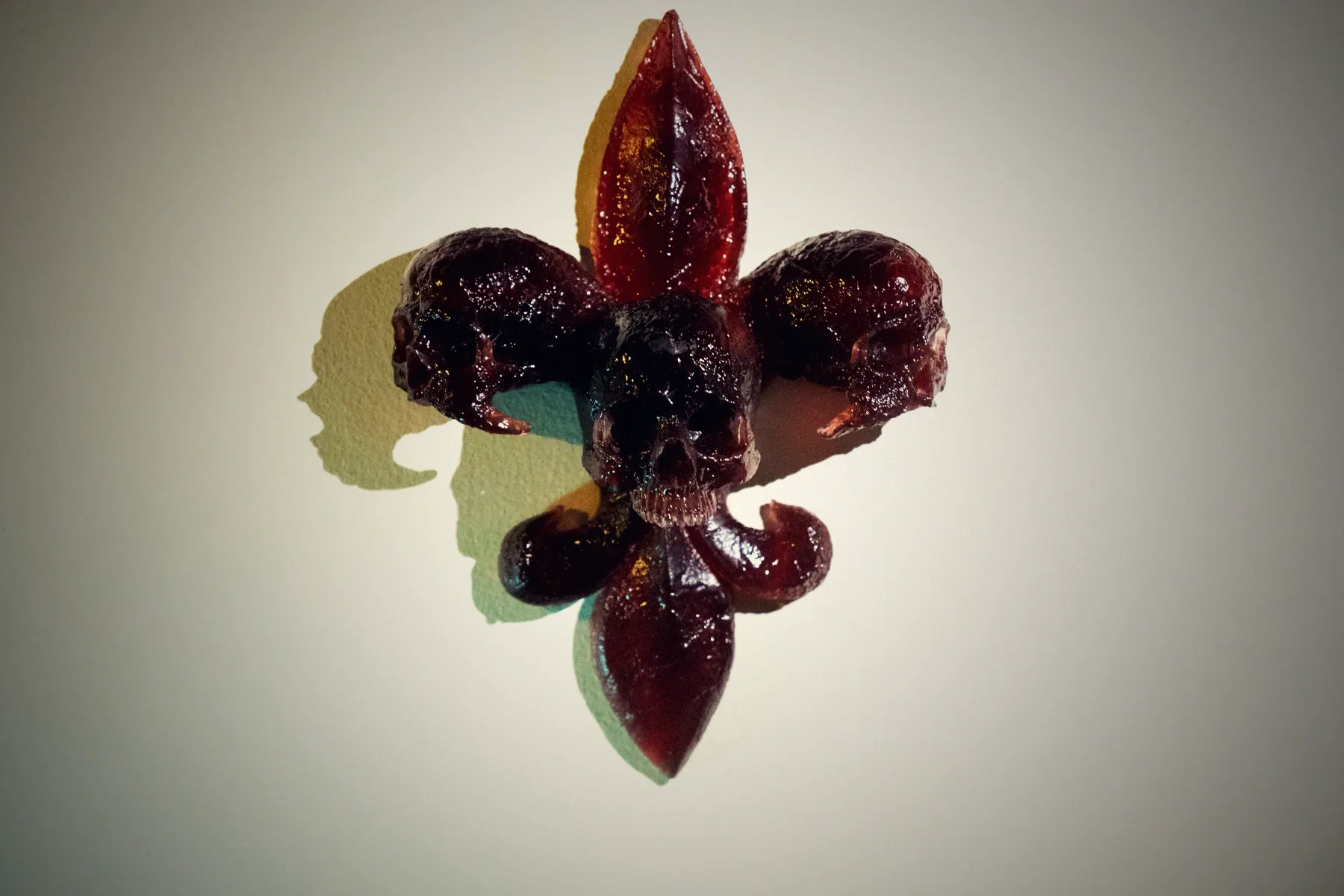 A fleur-de-lis built from skulls, cast in deep red translucent resin, mounted on a pale wall with its shadow beside it